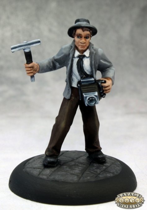Figurine of a person holding a flash unit and camera on a gray background.  Reaper Miniatures Lacy O'Malley muckraker painted